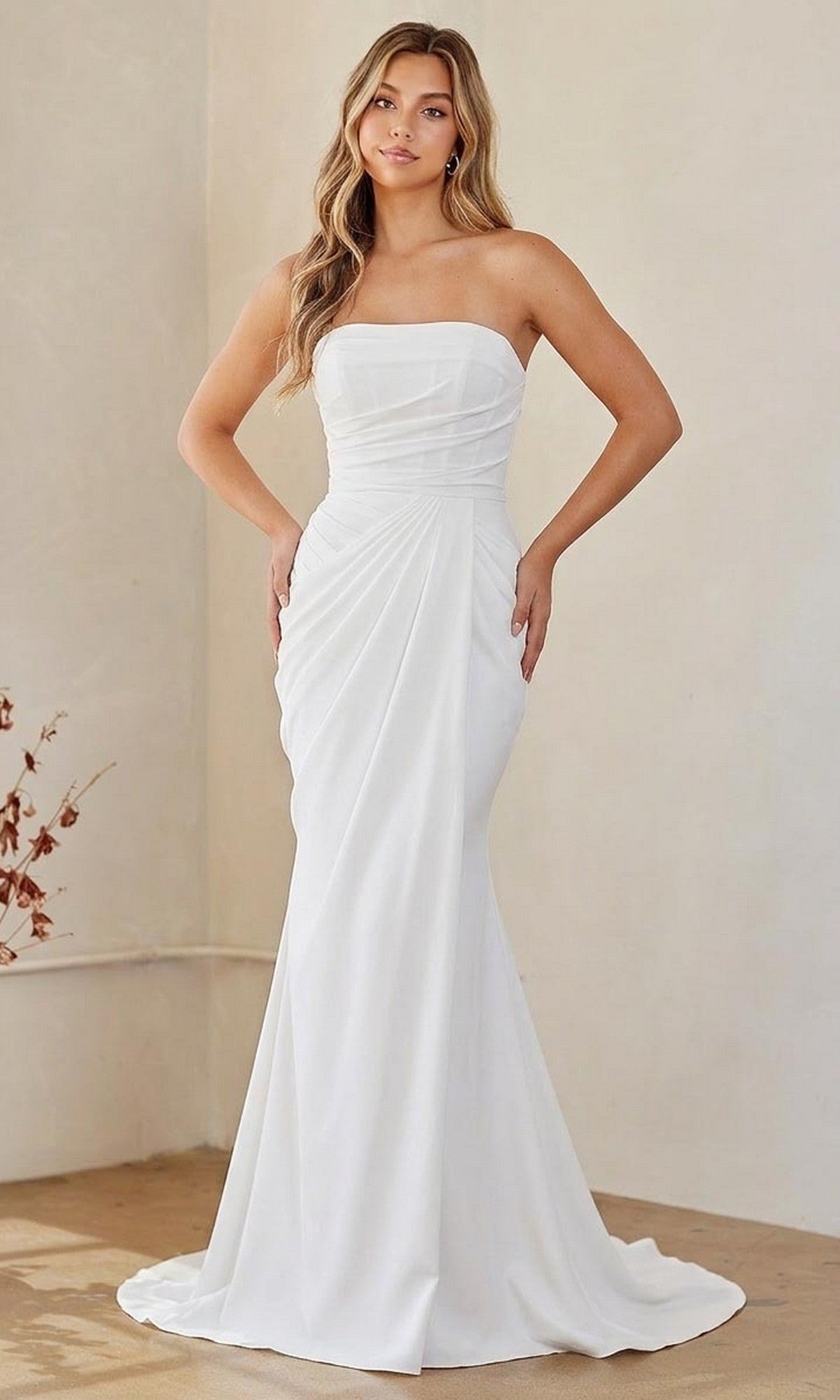 A woman stands indoors wearing the Long Formal Dress: Amelia Couture 6149 in white, a strapless evening gown with fitted silhouette and drape detail, her hands elegantly resting on her hips.